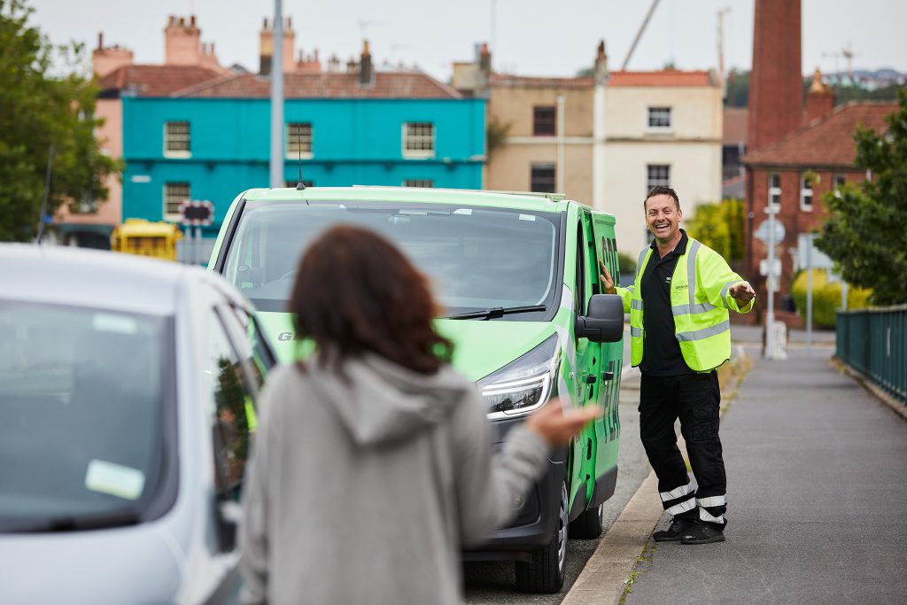 EV technician next to a Green Flag van buzz-ing to see a customer, both waving at each other.