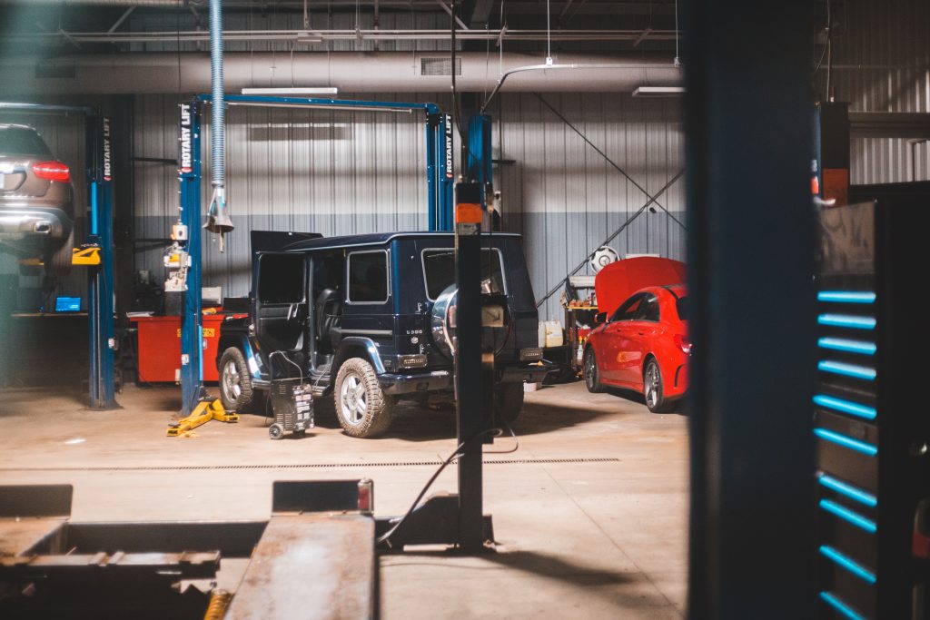 three cars in a repair workshop. left one is lifted in air only partly in the picture, middle is a midnight blue jeep with a door open, right is a red saloon-type car with the bonnet popped also partly out of frame.