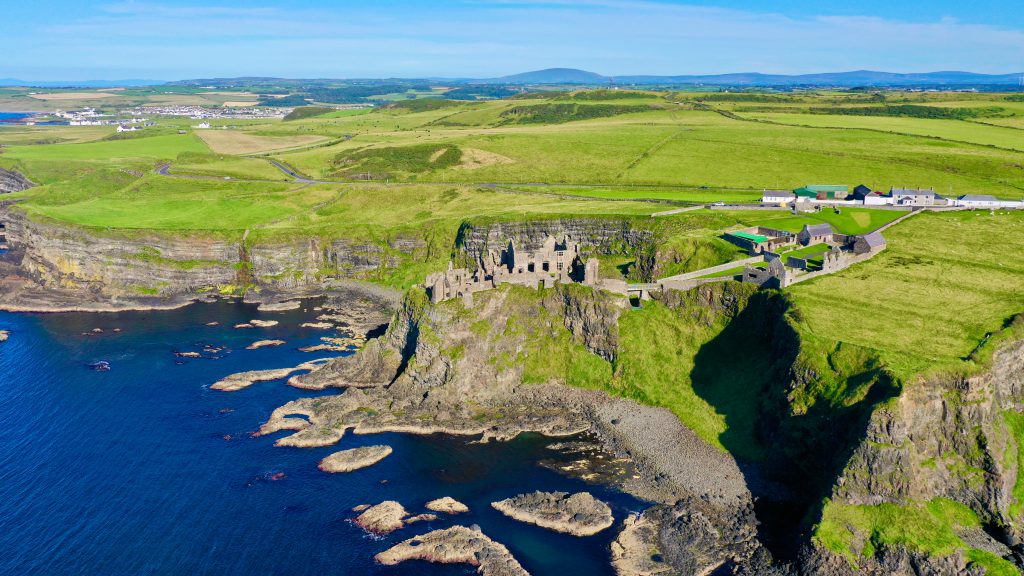 aerial shot of Dunluce Castle, listed first in the list of places to visit on the road trip in Northern Ireland. With the deep blue sea in the bottom left of the frame, the castle in the middle, and green land filling the rest of the shot.