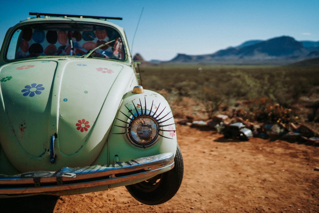 light green beetle with flowers painted onto the bonnet is parked on a dirt track. pov is a close up of the right side if the bonnet and light, which has eyelashes attached.