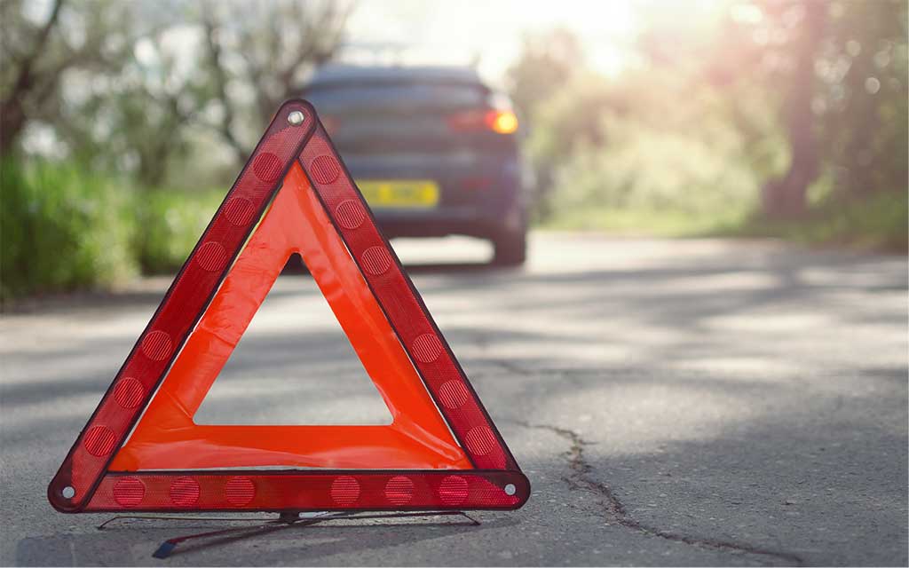 A blue car is stationary with its warning lights on. A warning triangle has been placed down the road from the car to alert oncoming traffic.