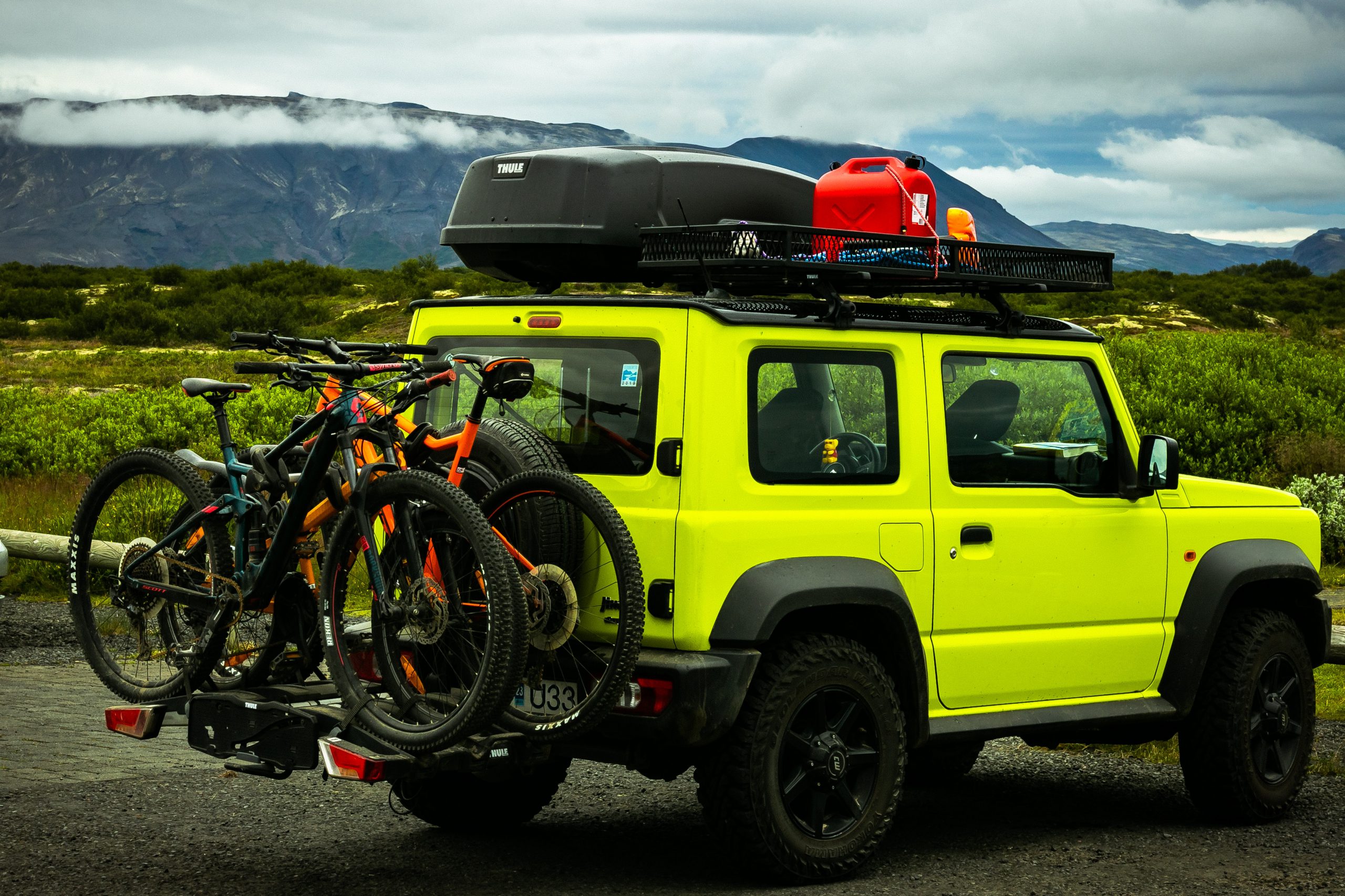 Yellow-ish green 4x4 with roof rack and bike rack attached. Luggage container and red box on roof rack and two bikes on bike rack. Greenery and mountains in the background.