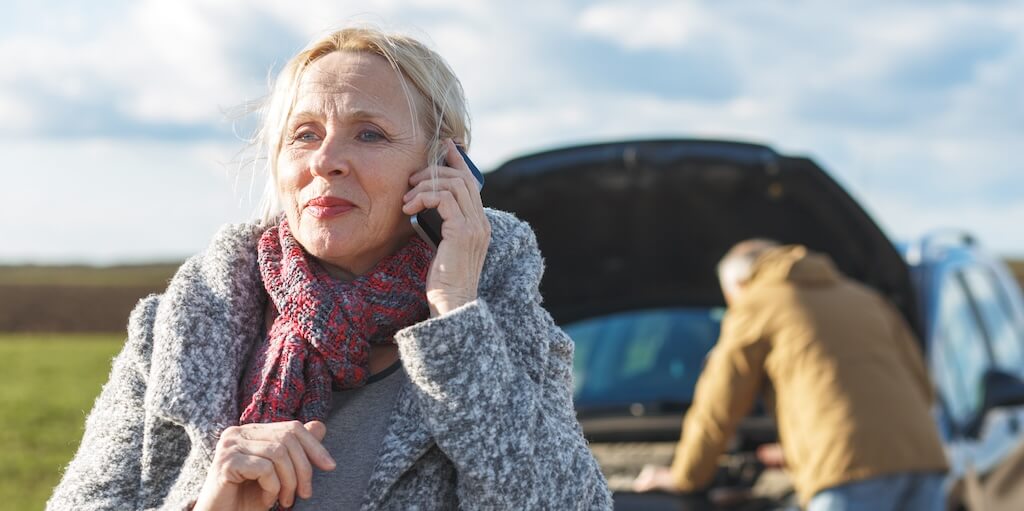 A lady in a coat and scarf is on the phone to her breakdown provider, while a man is seen behind her looking at the broken-down car.