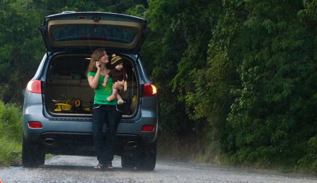 A woman and child are stood outside their car, calling their breakdown provider for help.
