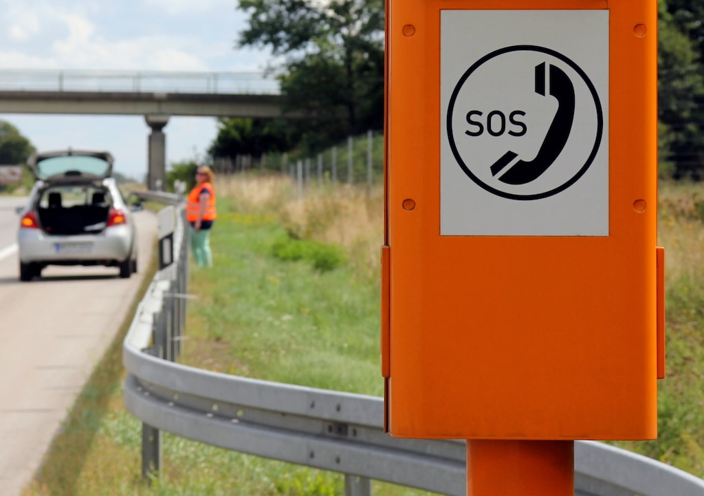 A driver waits behind the safety barrier at the side of a road. The driver is wearing a hi-vis vest. An orange SOS phone point is seen at the foreground of the image.