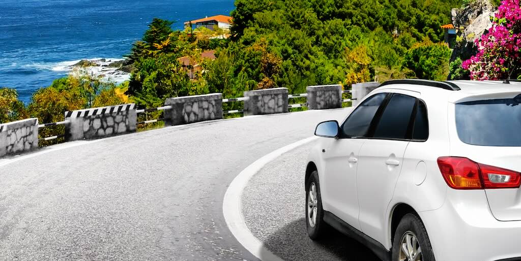 A white estate car turns a corner, overlooking the sea.
